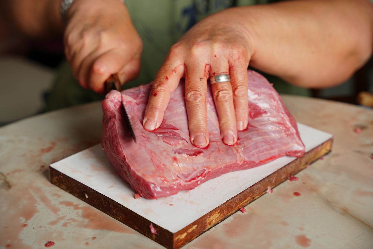 A detailed image of hands with a knife cutting a piece of raw meat on a cutting board indoors.